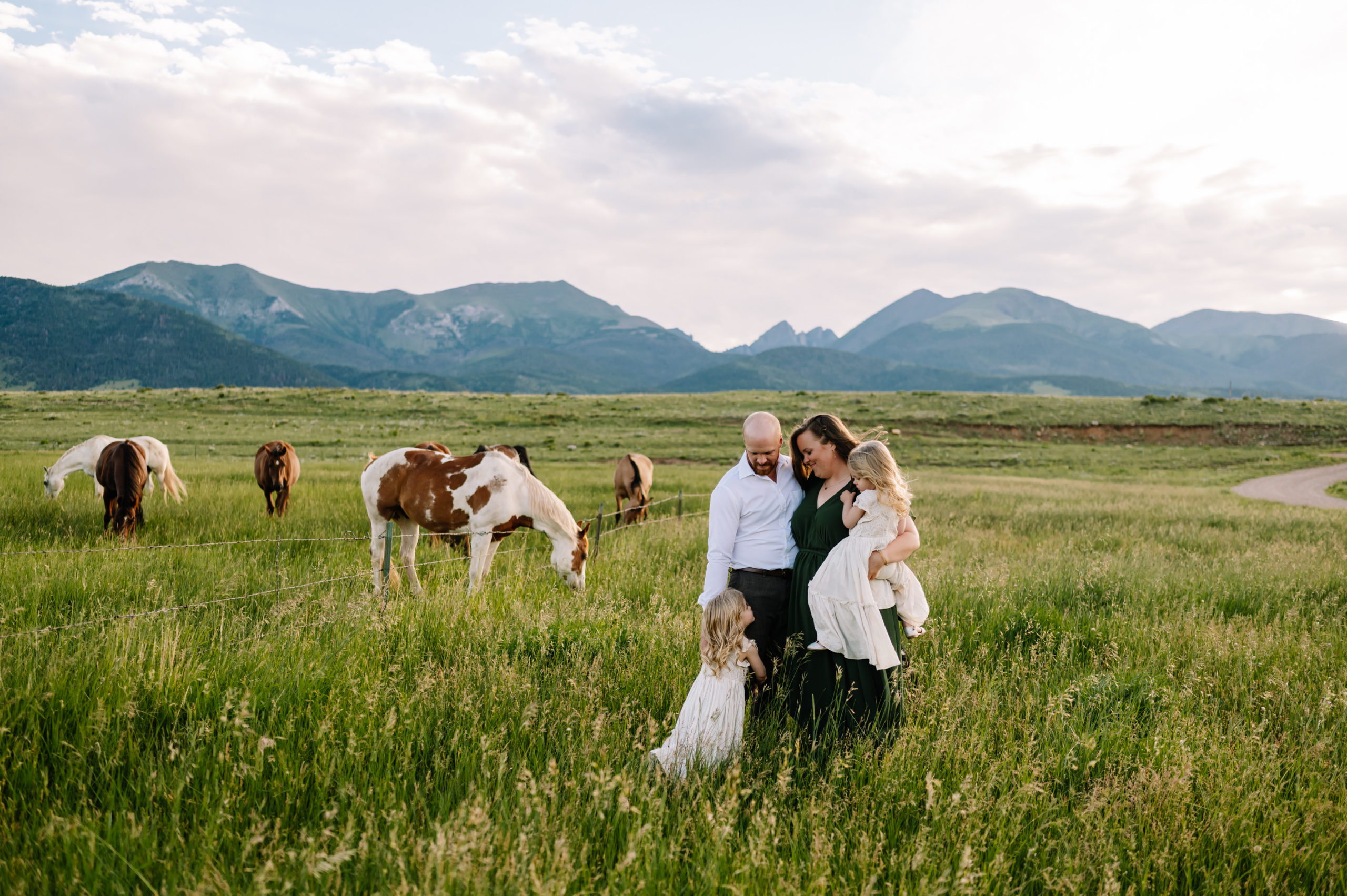 A mom, dad, and two twin girls hugging in a field of horses in front of the mountains.